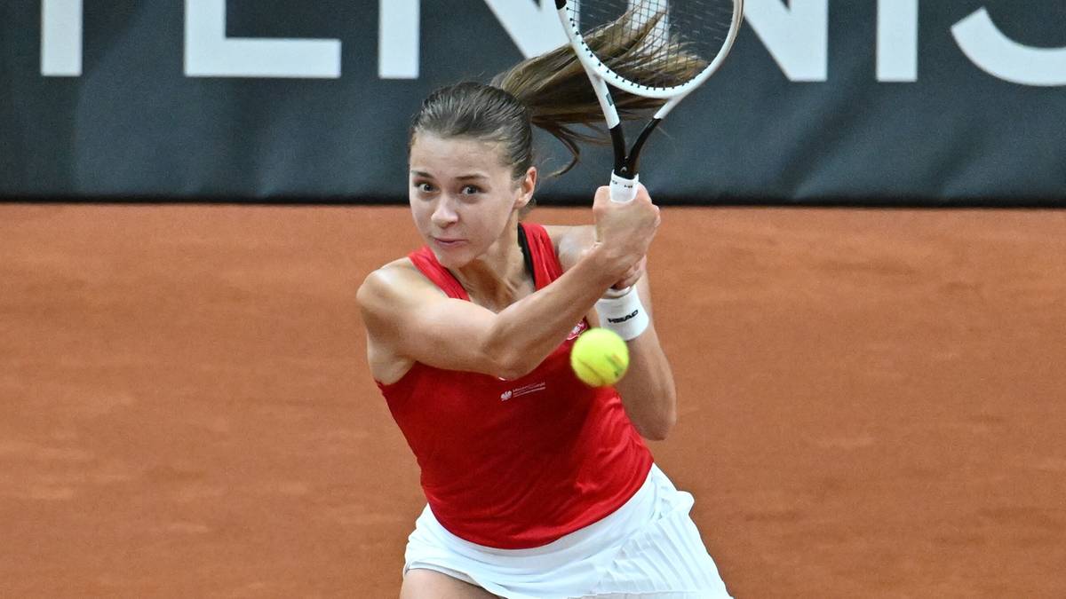 Tenisistka w czerwonej bluzce i białej sp&oacute;dniczce odbija piłkę rakietą podczas meczu na ceglanym korcie.