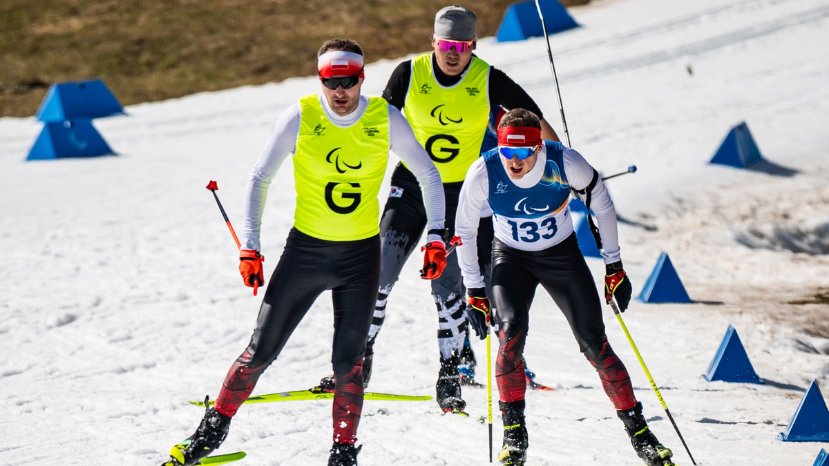 Trzech narciarzy w kombinezonach sportowych jedzie na nartach po ośnieżonej trasie, każdy z kijami narciarskimi. Dwóch z nich nosi kamizelki z logo Para sport. W tle widoczne są niebieskie pachołki.