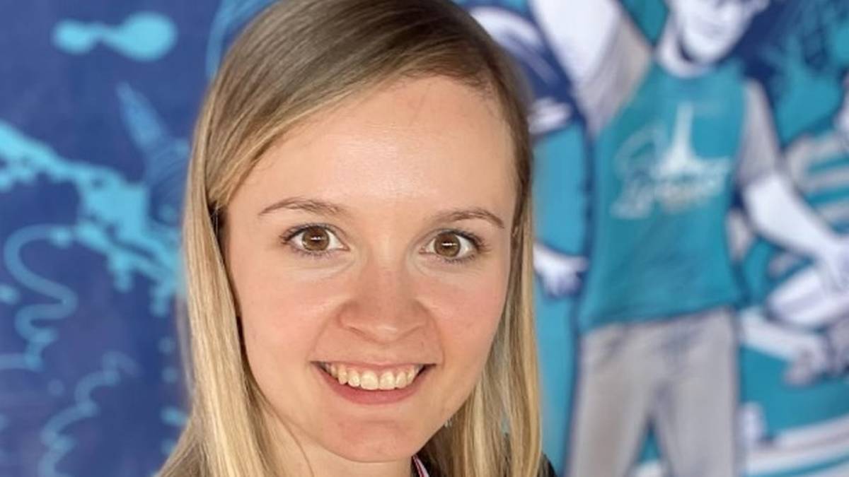 Kobieta z blond włosami i uśmiechem na twarzy, stojąca na tle niebieskiej ściany z grafiką przedstawiającą rolkarzy.