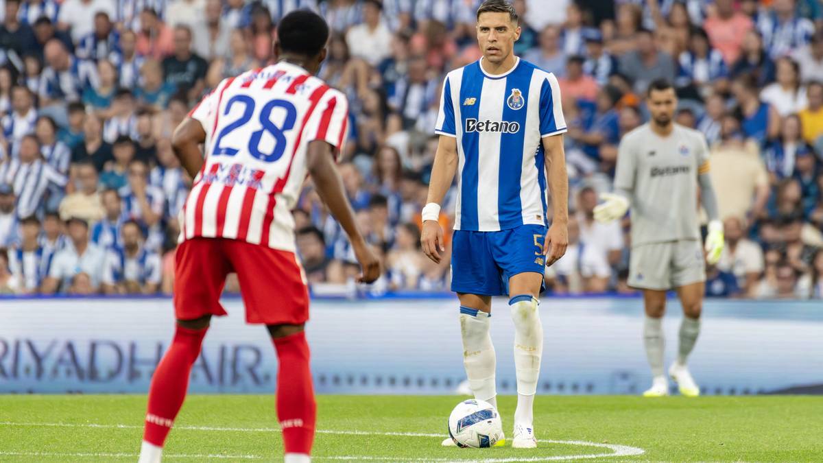 Piłkarz w białej koszulce z czerwonymi pasami i numerem 28 na plecach stoi tyłem do widza, zwracając się w stronę piłkarza w niebiesko-białej koszulce z logo FC Porto, który przygotowuje się do wykonania rzutu wolnego. W tle widoczni są inni zawodnicy i liczni kibice na trybunach.