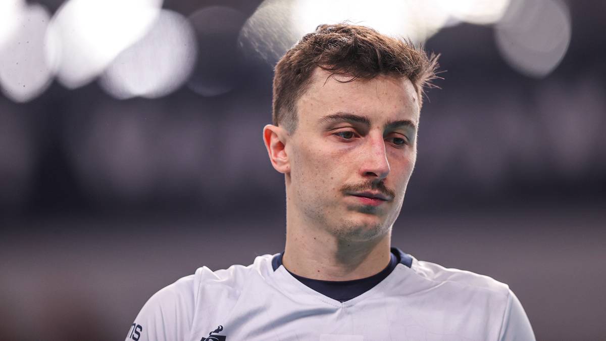 Zbliżenie na twarz młodego siatkarza, mężczyzny o jasnej karnacji i ciemnych włosach, z wąsami. Jest ubrany w biały str&oacute;j sportowy.