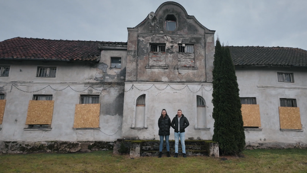 Para stoi przed zniszczonym budynkiem z zabitymi oknami