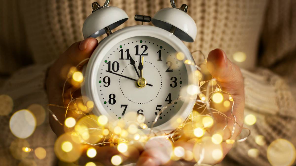 Biały budzik z dwiema dzwonkami, trzymany w dłoniach otoczonych girlandą światełek.