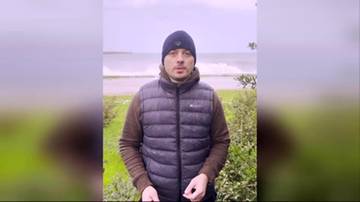 Mężczyzna w zimowej czapce i puchowej kamizelce stoi na tle krajobrazu z morzem w tle.