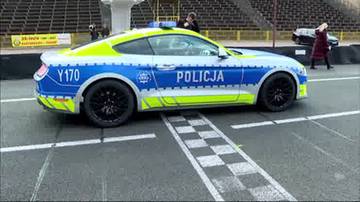 Samoch&oacute;d policyjny Ford Mustang w jaskrawej kolorystyce z napisem POLICJA, oznakowaniem Y170 i niebieskimi światłami na dachu, stoi na pasie startowym z liniami toru wyścigowego.