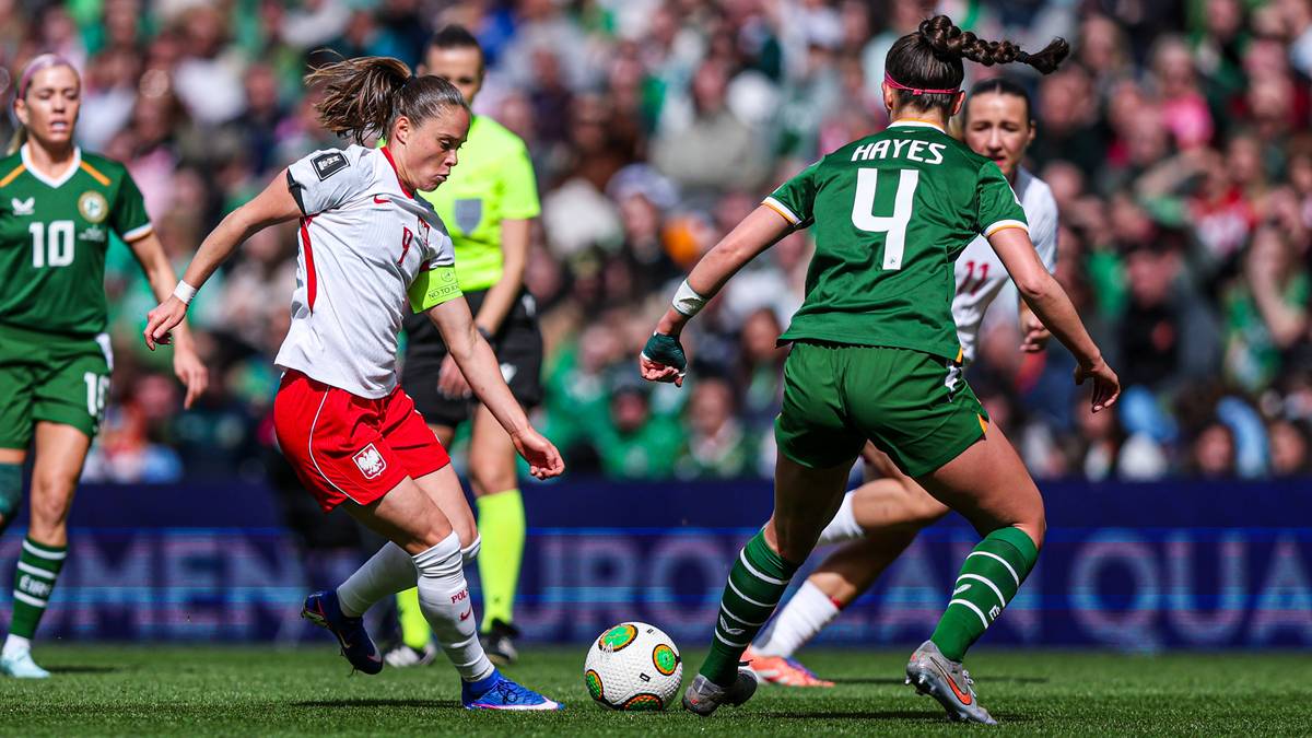 Piłkarka w białej koszulce z czerwonymi elementami pr&oacute;buje odebrać piłkę zawodniczce w zielonej koszulce.