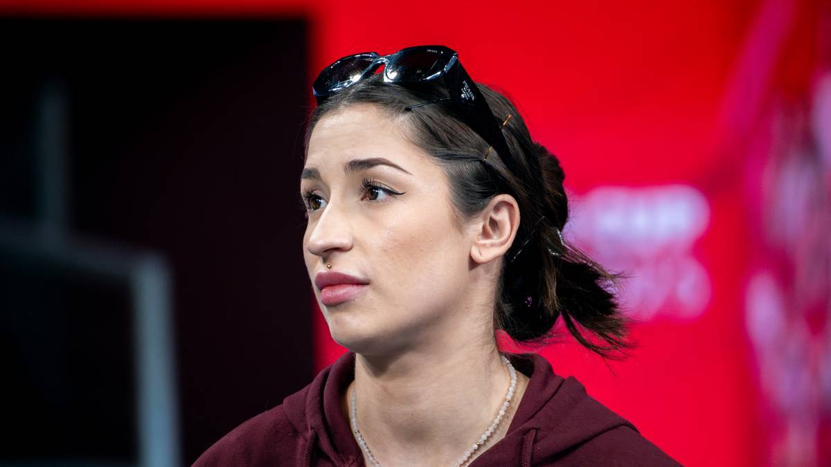 Portret młodej kobiety z ciemnymi włosami upiętymi do tyłu, w okularach przeciwsłonecznych na czole, na czerwonym tle.