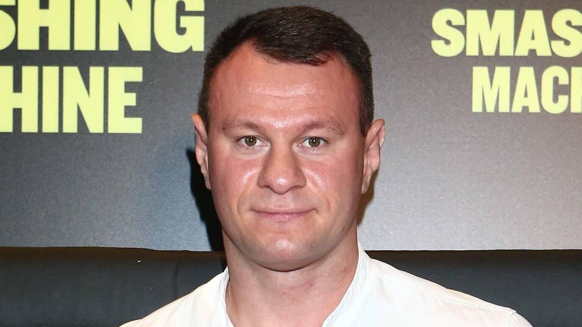 Mężczyzna w białej koszuli i jeansach stoi przed czarnym tłem z ż&oacute;łtym napisem "SMASHING MACHINE".