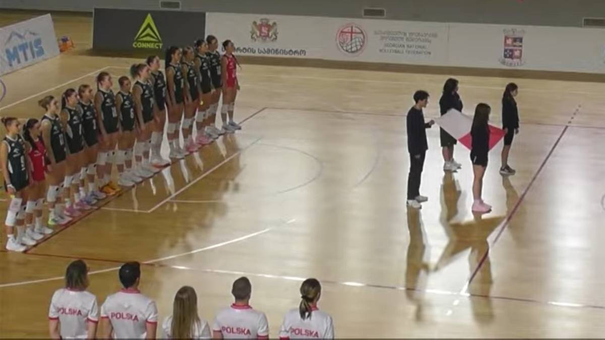 Drużyna siatkarek w czarnych strojach stoi na hali sportowej, a cztery osoby prezentują polską flagę.