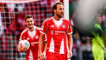 Dw&oacute;ch piłkarzy w czerwonych koszulkach z białymi pasami i logo "Magenta TV" i "Swarovski Optik". Jeden piłkarz po lewej trzyma piłkę w dłoni, drugi po prawej stoi z rękami opartymi na biodrach.