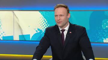 Mężczyzna w ciemnym garniturze, białej koszuli i bordowym krawacie, z polską flagą w klapie marynarki, stoi na tle ekranu telewizyjnego z abstrakcyjnymi grafikami.