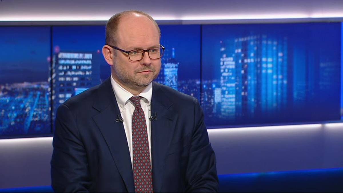 Mężczyzna w okularach i ciemnym garniturze siedzi na tle niebieskich ekran&oacute;w z widokiem na nocne miasto.