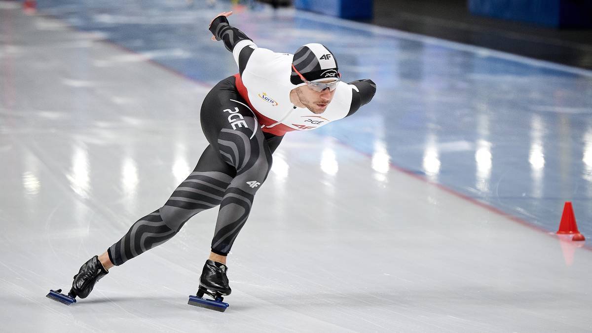 Łyżwiarz szybki w białym i czarnym stroju sportowym pochyla się podczas jazdy na lodowisku.
