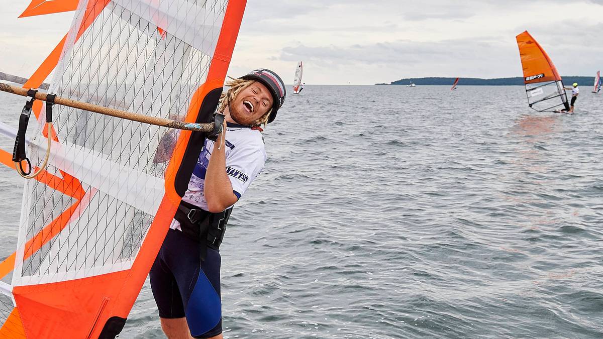 Młody mężczyzna w kasku i okularach pływa na desce windsurfingowej, trzymając bom. Uśmiecha się do obiektywu.