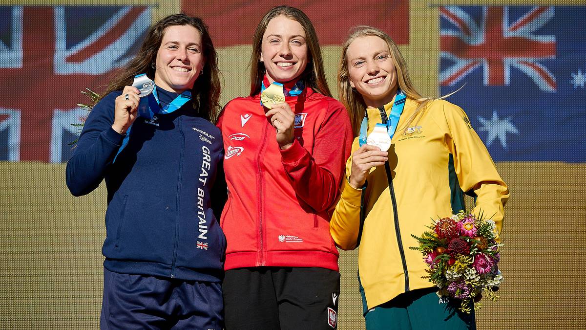 Trzy uśmiechnięte kobiety stoją na podium, trzymając medale i dyplomy. Na ich szyjach wiszą medaliony, a w rękach trzymają bukiety kwiatów. Kobieta po lewej stronie ubrana jest w granatową bluzę z napisem "GREAT BRITAIN". Kobieta w środku ma na sobie czerwoną kurtkę. Kobieta po prawej stronie ma na sobie żółtą kurtkę.