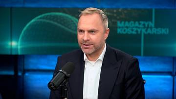 Łukasz Koszarek, ubrany w garnitur, udziela wywiadu przed mikrofonem na tle planszy "Magazyn Koszykarski".