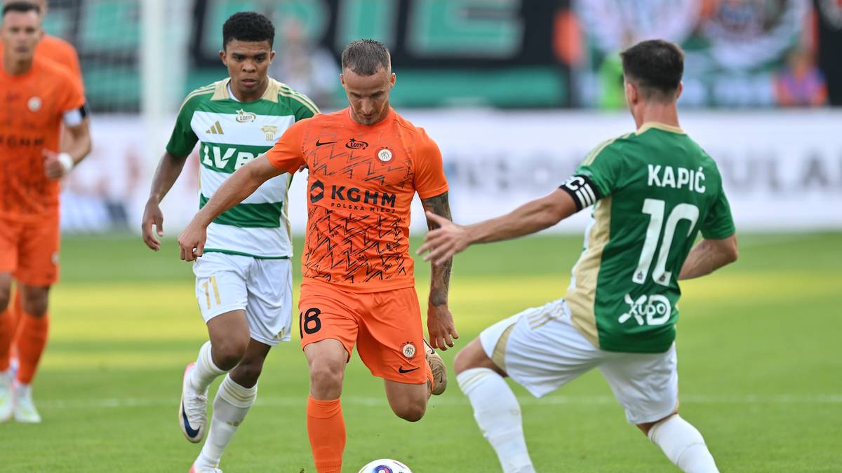Piłkarze w pomarańczowych i zielonych strojach walczą o piłkę na boisku podczas meczu PKO BP Ekstraklasy.