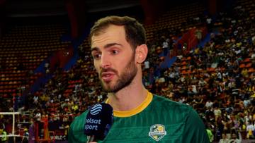 Mężczyzna w zielonej koszulce z żółtym kołnierzykiem, z krótką brodą i włosami, trzymający mikrofon przed ustami. W tle widać trybuny wypełnione kibicami.