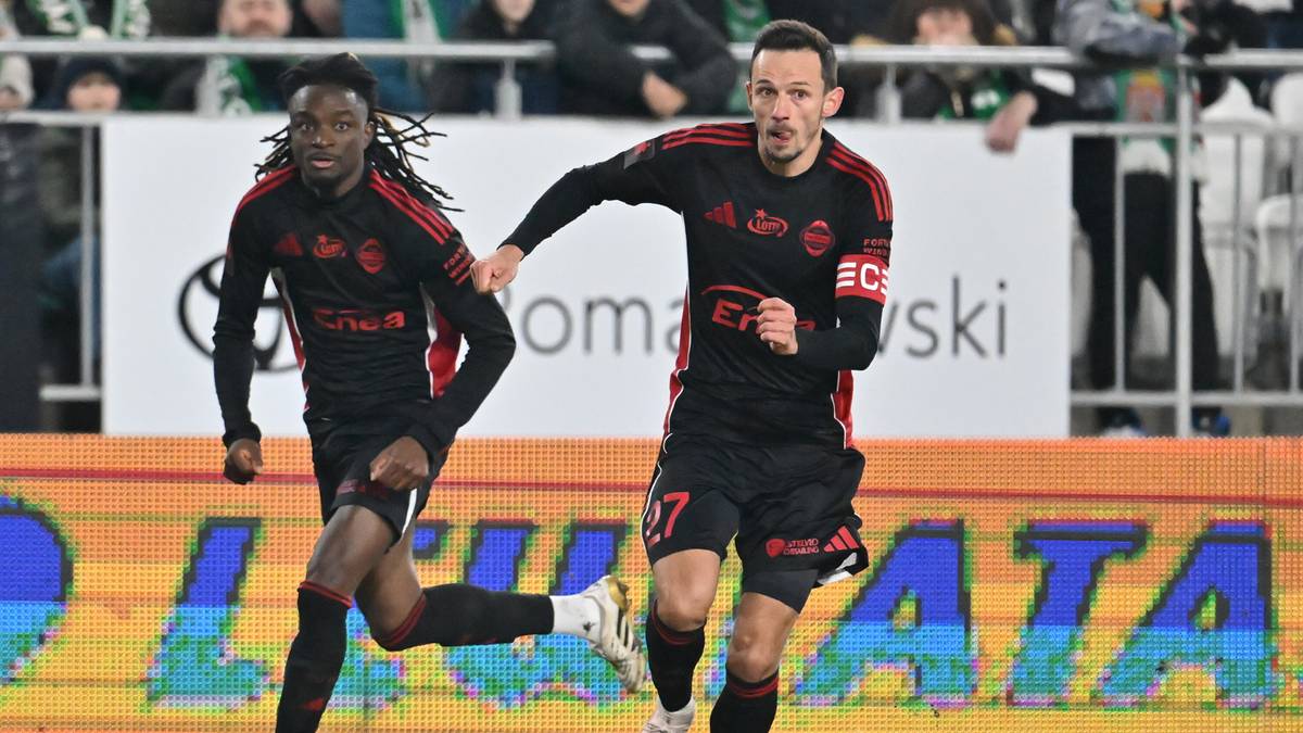 Dw&oacute;ch piłkarzy w czarnych strojach z czerwonymi elementami biegnie po boisku z piłką nożną.