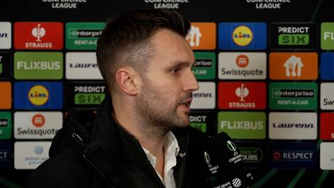 Mężczyzna w średnim wieku o kr&oacute;tkich włosach i z zarostem, m&oacute;wiący do mikrofonu na tle ścianki z logo sponsor&oacute;w.