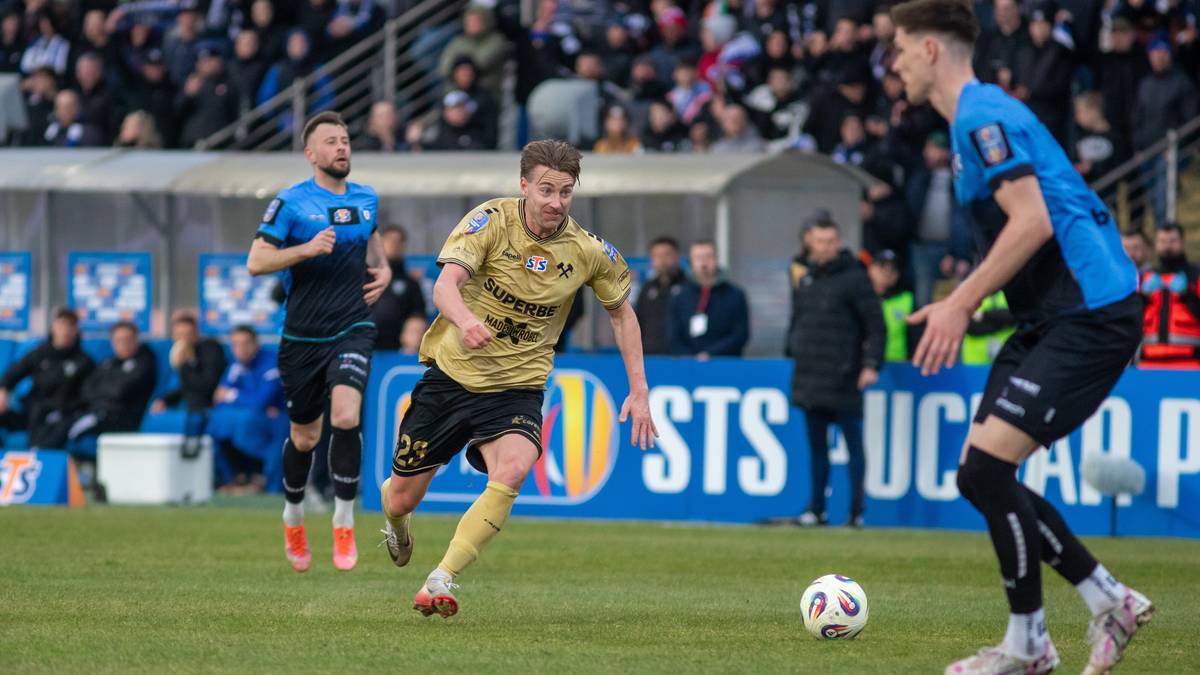 Dw&oacute;ch zawodnik&oacute;w piłki nożnej rywalizuje o piłkę na zielonym boisku stadionu, w tle widownia.