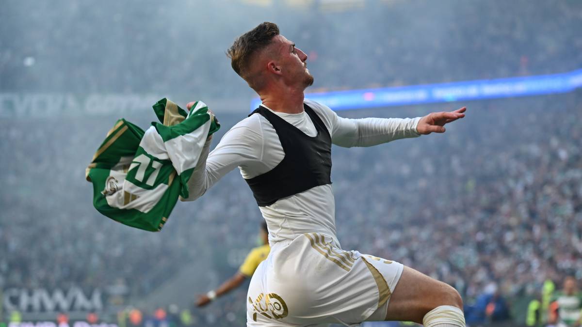 Piłkarz Lechi Gdańsk w białym stroju świętuje gol, trzymając w ręku zielono-białą koszulkę z numerem 77. Meczem odbywa się na stadionie, w tle widać trybuny wypełnione kibicami.