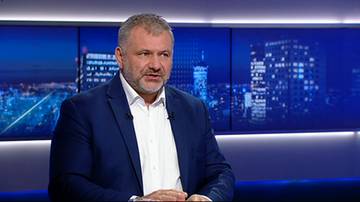 Mężczyzna w białej koszuli i granatowej marynarce siedzi i m&oacute;wi, na tle widać nocną panoramę miasta.