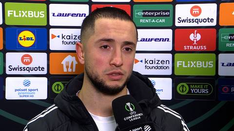 Portret mężczyzny z kr&oacute;tkimi włosami i zarostem, stojącego przed ścianą z logotypami sponsor&oacute;w.