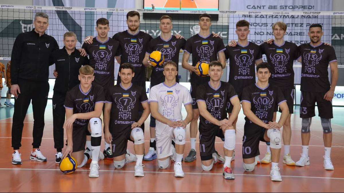 Drużyna siatkarska w komplecie, zawodnicy w czarnych strojach z fioletowymi elementami, pozują do zdjęcia na boisku siatkarskim.