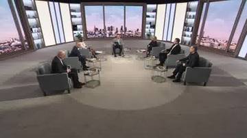 Grupa siedmiu mężczyzn w garniturach dyskutuje w studiu telewizyjnym.