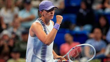 Iga Świątek unosi zaciśniętą pięść, trzymając rakietę tenisową, w tle widzowie na trybunach.