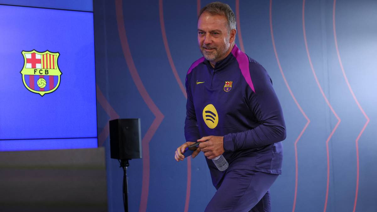 Trener Hansi Flick w dresie FC Barcelony, stojący przed niebieską ścianą z herbem klubu.