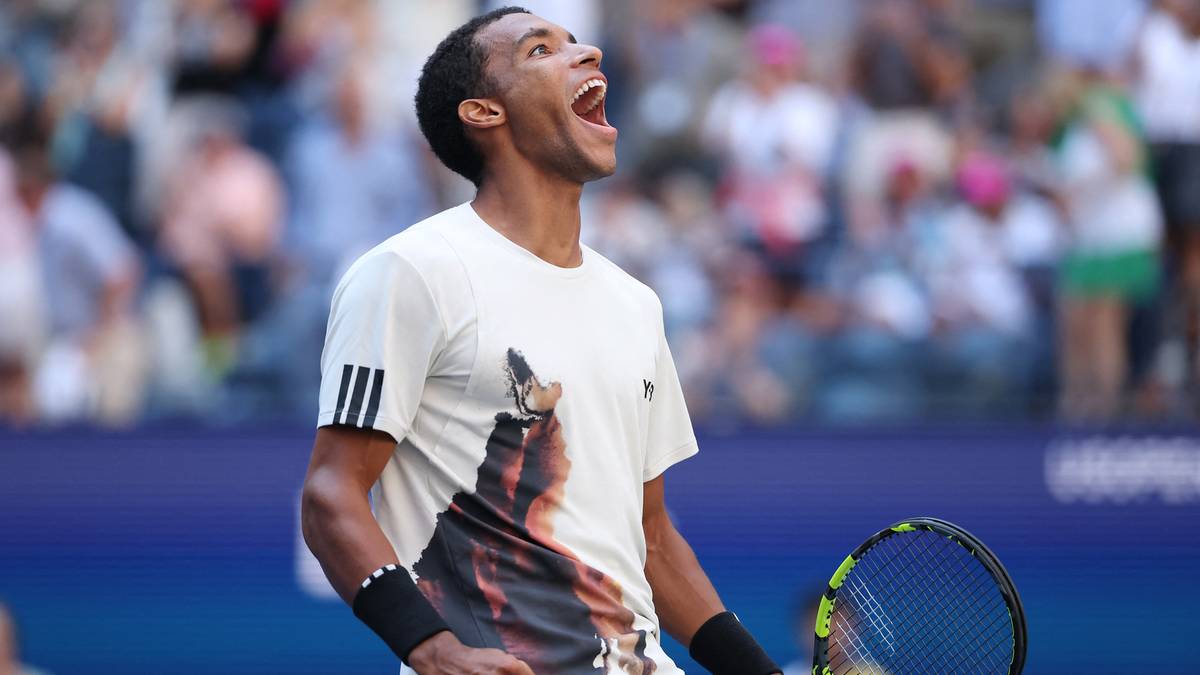 Zawodnik tenisa w bieli z krzykiem radości, po wygranej piłce. Widoczne jego ramiona, szyja, twarz i część rakiety. W tle widzowie na trybunach.