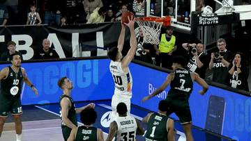 Koszykarz w białej koszulce z numerem 10 wykonuje rzut do kosza, otoczony innymi zawodnikami.