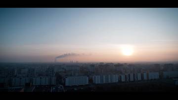 Panorama miasta o wschodzie lub zachodzie słońca, z widocznym słońcem, niebem w odcieniach błękitu i r&oacute;żu oraz dymem unoszącym się z fabrycznych komin&oacute;w w oddali.