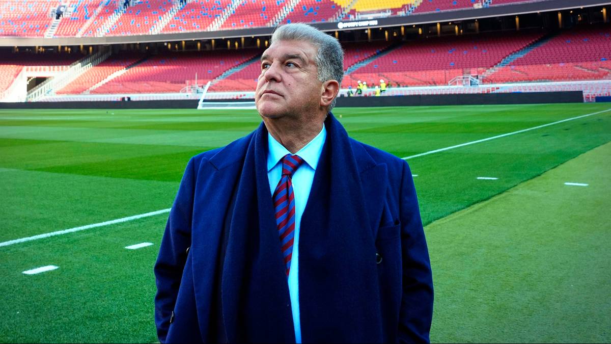 Portret starszego mężczyzny w granatowym płaszczu i krawacie, stojącego na tle zielonej murawy stadionu piłkarskiego.