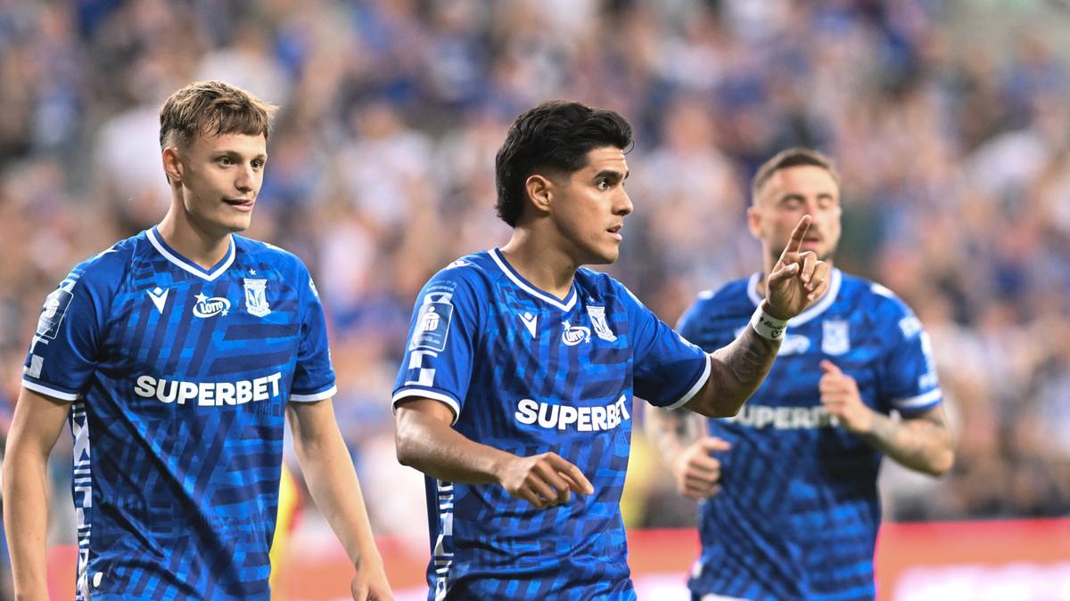 Trzech piłkarzy w niebieskich strojach sportowych z białymi napisami "SUPERBET" na piersi i niebieskimi herbami klubowymi na rękawach. Gracz po lewej patrzy w bok, środkowy jest odwrócony lekko w prawo i pokazuje palcem, a gracz po prawej biegnie i też patrzy w bok. W tle widać rozmazanych kibiców na trybunach.