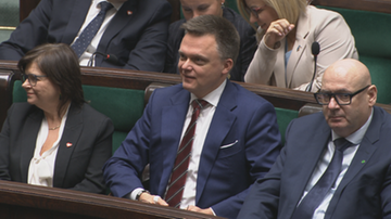 Mężczyzna w garniturze i krawacie uśmiecha się, siedząc w ławach sejmowych obok innych osób.