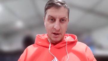 Portret mężczyzny w pomarańczowej bluzie z kapturem i białymi słuchawkami, patrzącego prosto w obiektyw.