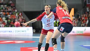 Dwie zawodniczki grające w piłkę ręczną. Jedna z nich, w białej koszulce z numerem 22, posiada czerwoną piłkę i przygotowuje się do podania. Druga zawodniczka, w czerwonej koszulce z numerem 14, blokuje ją.