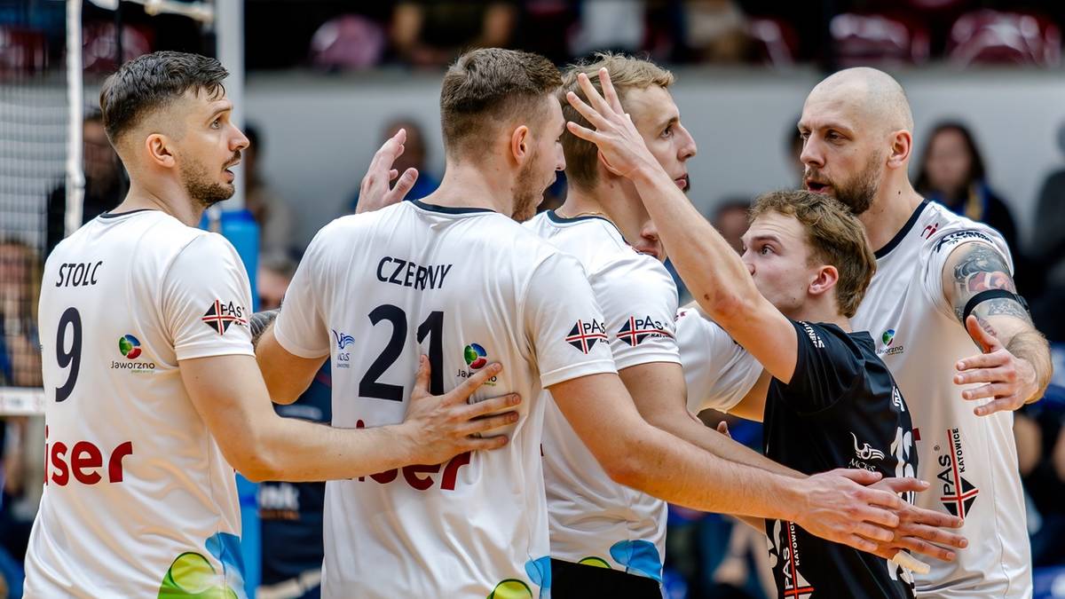 Sześciu siatkarzy w białych koszulkach z numerami i napisami na plecach, stojących blisko siebie na boisku siatkarskim, rozmawiających i gestykulujących.