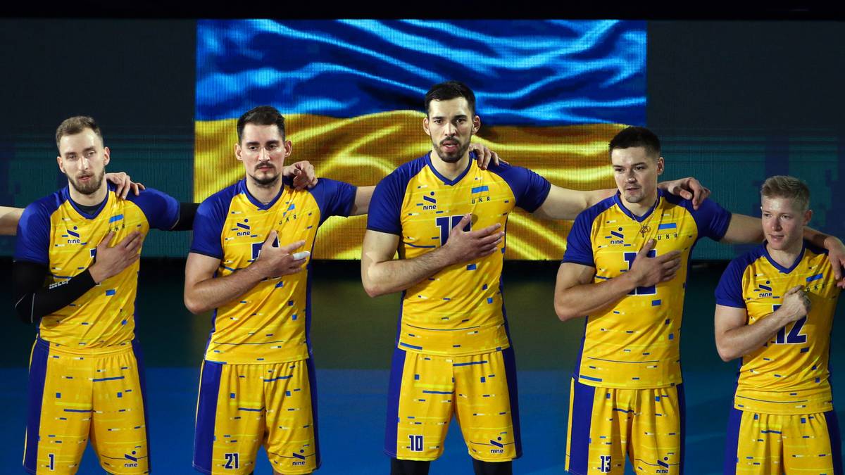 Pięciu siatkarzy reprezentacji Ukrainy w żółto-niebieskich strojach stoi przed flagą narodową z rękami na sercu.