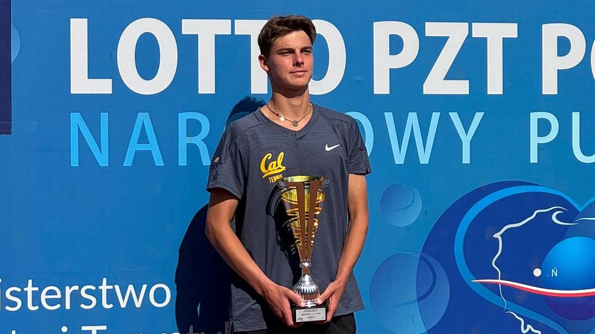 Młody mężczyzna w koszulce tenisowej z pucharem w rękach, na tle niebieskiego banera.