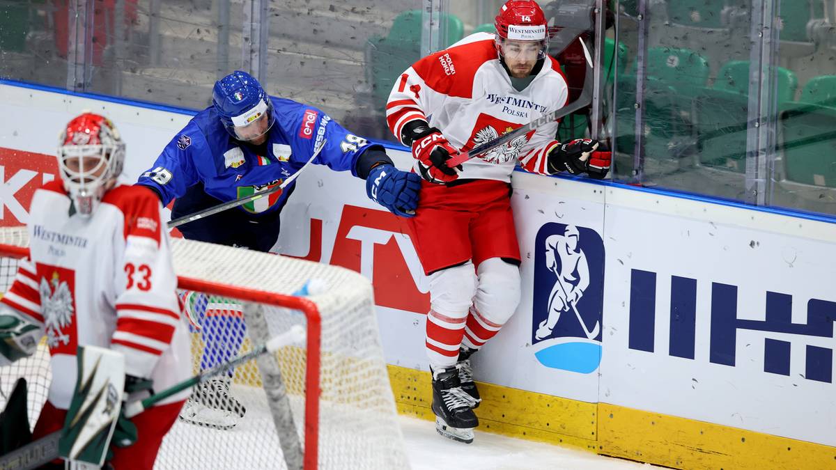 Dwóch zawodników hokejowych walczy o krążek przy bandzie, bramkarz w bramce