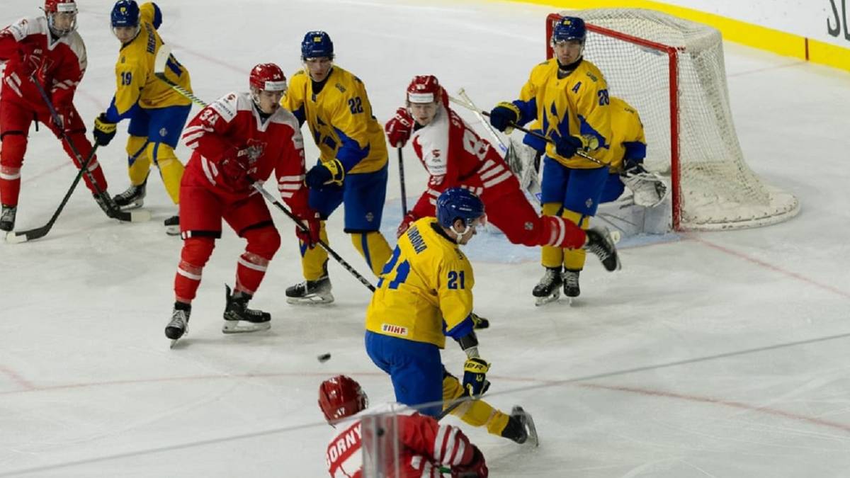 Drużyna hokejowa w czerwonych strojach walczy z drużyną w żółtych strojach. Jeden z graczy w czerwonym stroju leży na lodzie, próbując złapać krążek, podczas gdy zawodnicy w żółtych strojach próbują go odebrać.