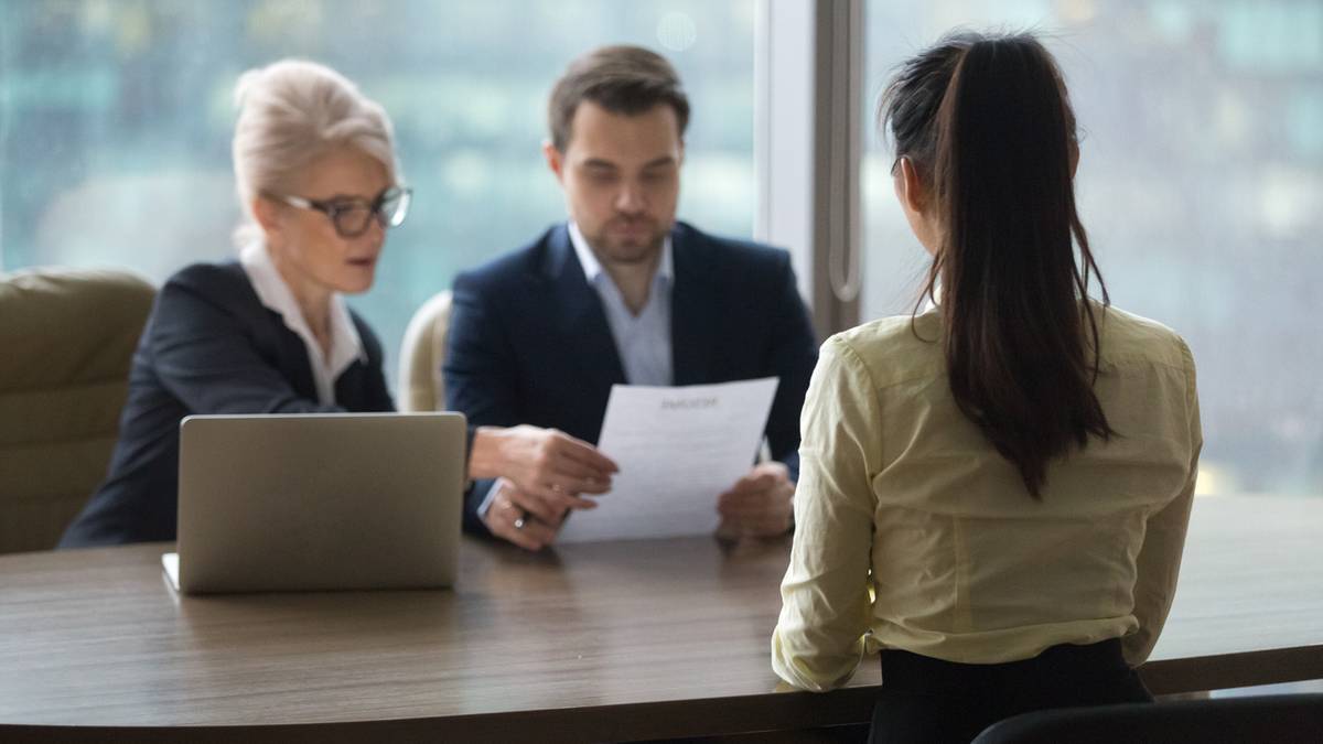 Trzy osoby siedzące przy biurku podczas rozmowy kwalifikacyjnej, dw&oacute;ch rekruter&oacute;w analizuje CV, a kandydatka odpowiada.