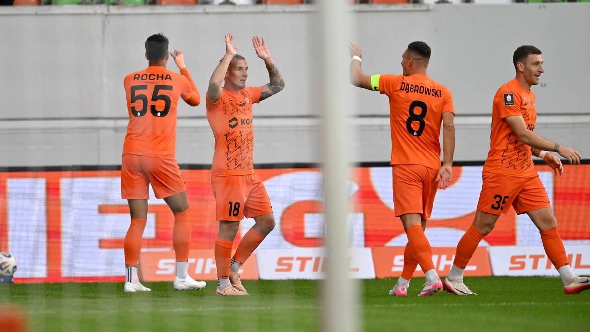 Czterech piłkarzy w pomarańczowych strojach świętuje na boisku piłkarskim. Dwóch z nich unosi ręce w geście radości, a dwaj pozostali mają ręce na biodrach, uśmiechając się.