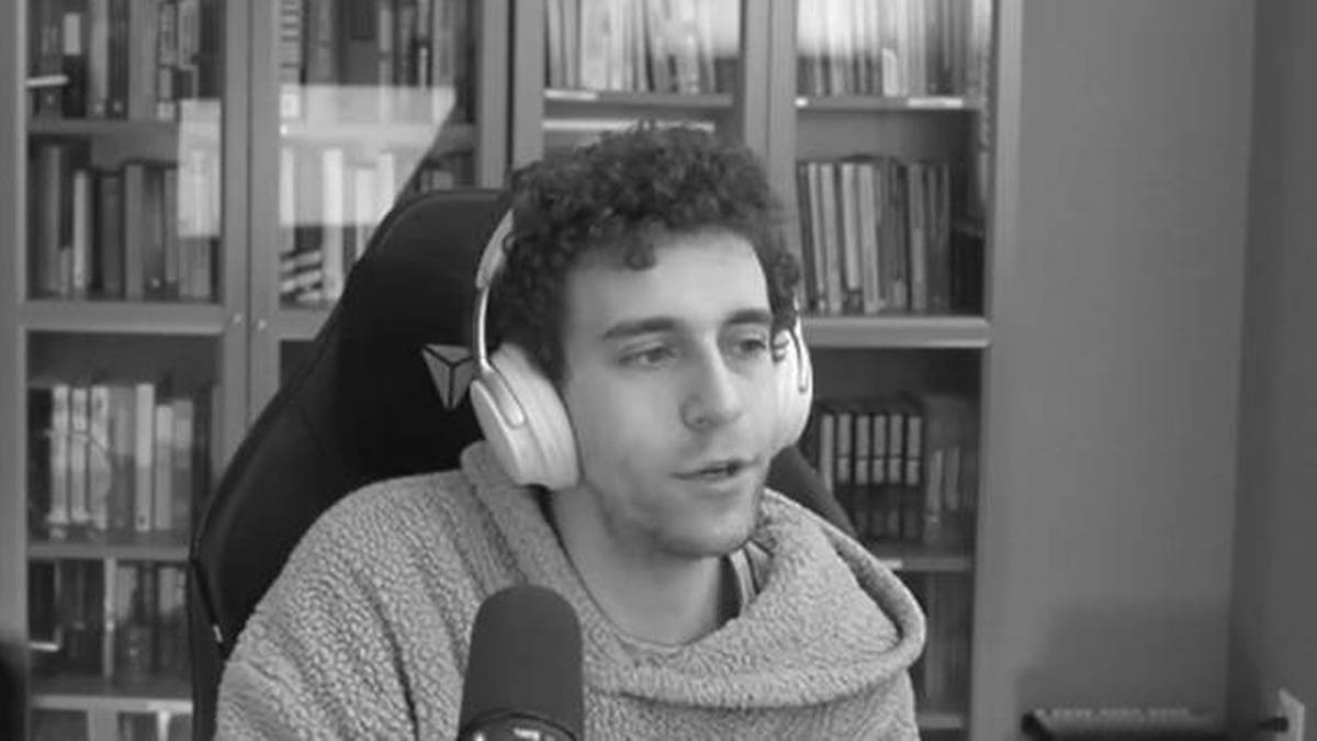 Młody mężczyzna z kręconymi włosami, noszący słuchawki, siedzi przed mikrofonem i patrzy na rozmówcę. Za nim znajdują się regały z książkami.