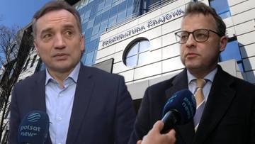 Dwaj mężczyźni w garniturach stoją przed budynkiem Prokuratury Krajowej, jeden z nich udziela wywiadu.