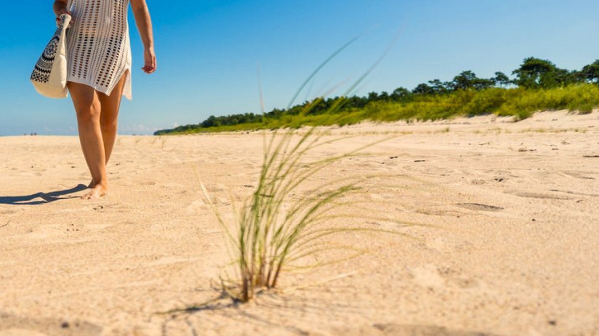 Kobieta w białej tunice idzie po piaszczystej plaży, trzymając w ręku torbę plażową. W tle widać zielone drzewa i błękitne niebo.